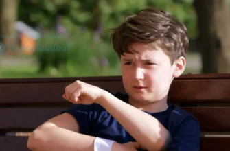 A young boy, around 7 years old, sitting on a park bench, gently holding his arm. His arm has a bandage on it. He has a slightly concerned but calm expression. Soft, natural lighting. Background shows a blurred park scene with trees and greenery.