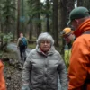 A realistic scene of a forest with a woman in her 60s or 70s, wearing warm clothing, standing near a path. She looks relieved and is surrounded by search and rescue personnel who appear to be assisting her. The forest is dense with trees, and there is a sense of relief and safety in the air.