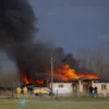A realistic photograph of a burning building in a rural area of Yaroslavl region, with emergency vehicles and firefighters visible, dark smoke rising into the sky, somber mood, no text or symbols