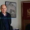 A realistic photograph of a man in his 30s wearing a plain dark jacket and jeans, standing in a modest office room with a Russian flag on the wall, his hands bound with rope, a serious expression on his face, soft natural lighting, high detail, no text or symbols on the image